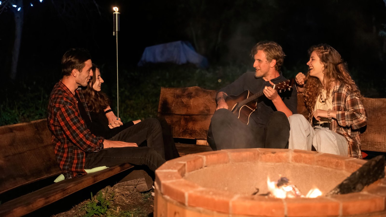 A group of happy young friends near a campfire at glamping, night. Two men and women. Playing guitar